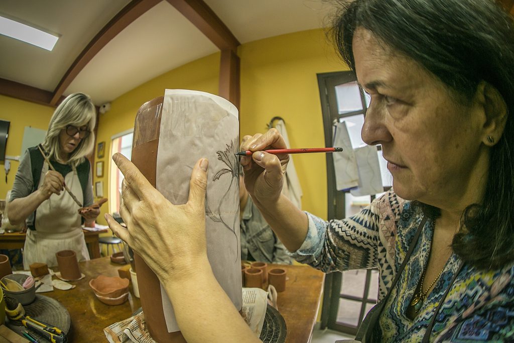 Foto de mulher desenhando em papel vegetal acima de objeto de cerâmica em laboratório com outras pessoas.