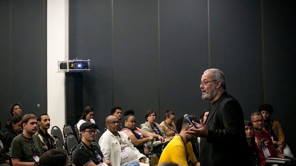 Foto com grupo de pessoas assistindo um homem dando palestra em auditório .