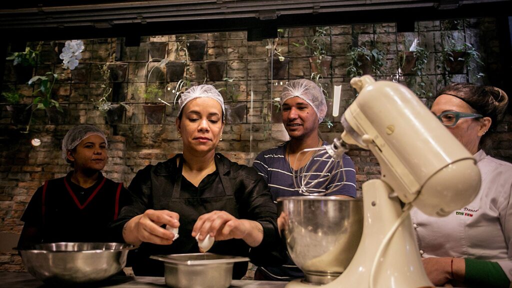 Foto de três pessoas assistindo como preparar bolo e uma delas quebrando ovos para bater na batedeira em cozinha profissional.